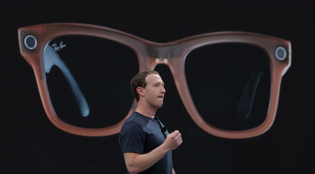 Mark Zuckerberg  fazendo uma palestra com uma imagem do óculos inteligente gigante ao fundo.